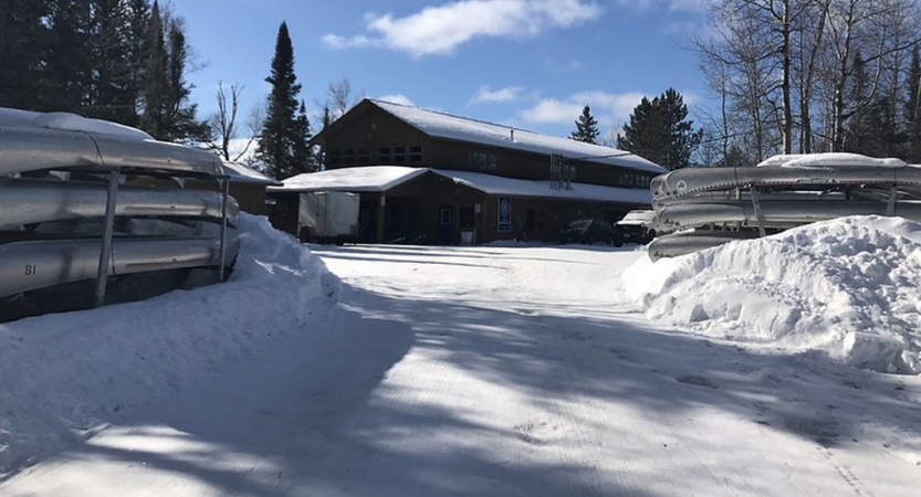 A building sits in a snowy landscape, with racks of canoes framing each side. 